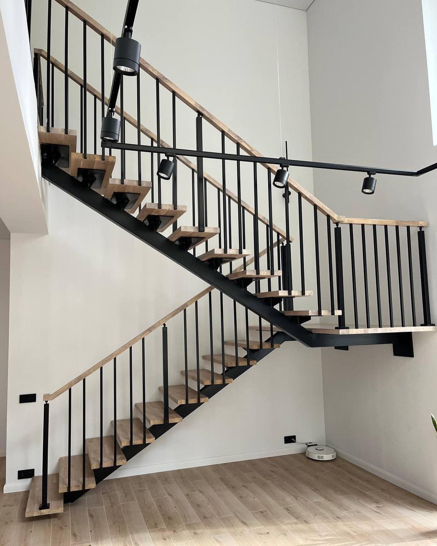 Modern wooden staircase with black metal railings and track lighting against a white wall.