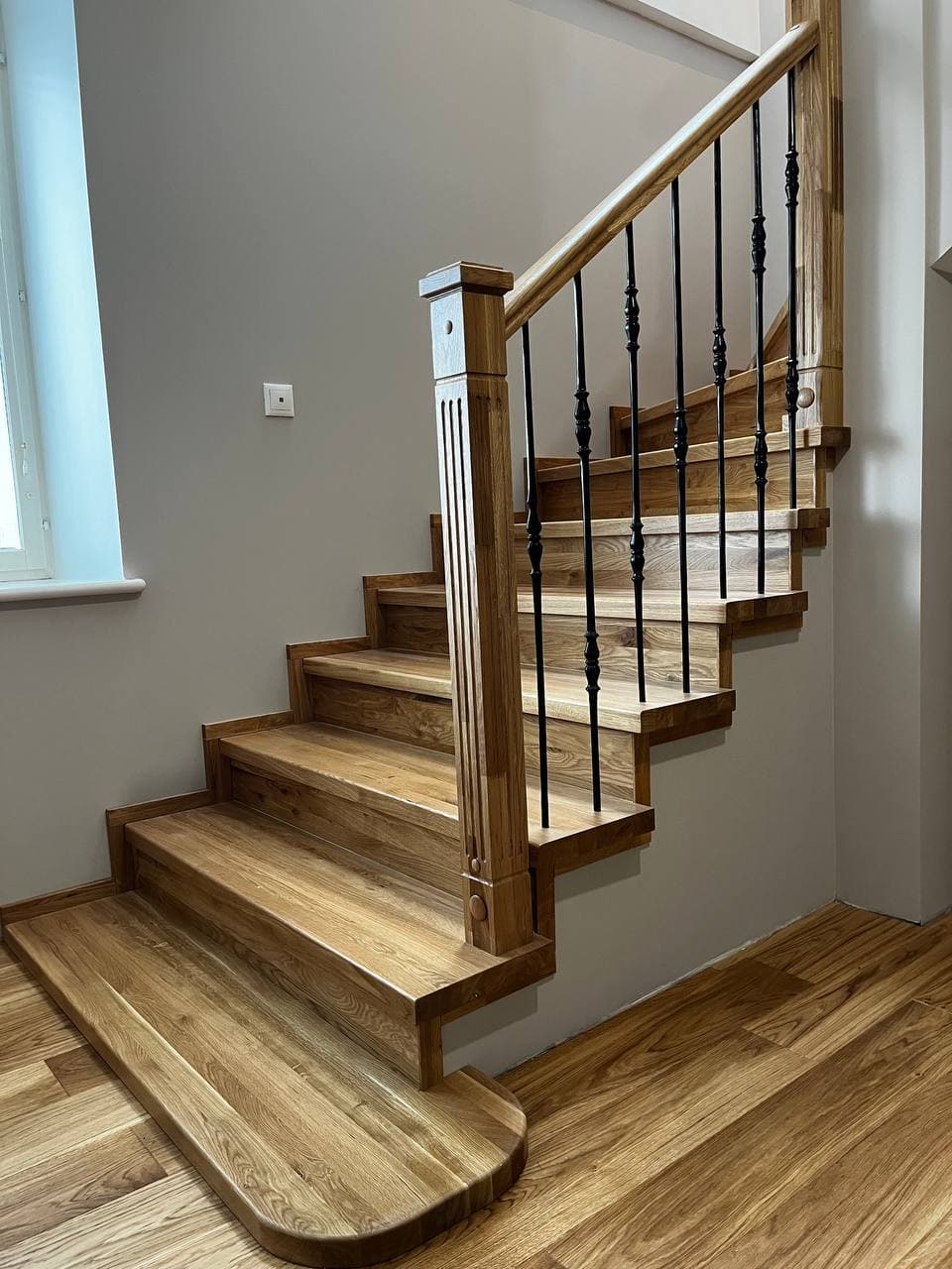 Light oak staircase with black metal spindles, a fluted newel post, and rounded bottom step.